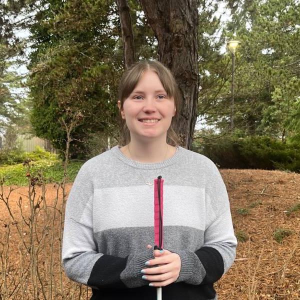 A headshot of Jordan. She smiles against a wooded background, wearing a gray and black striped sweater and holding a walking stick..